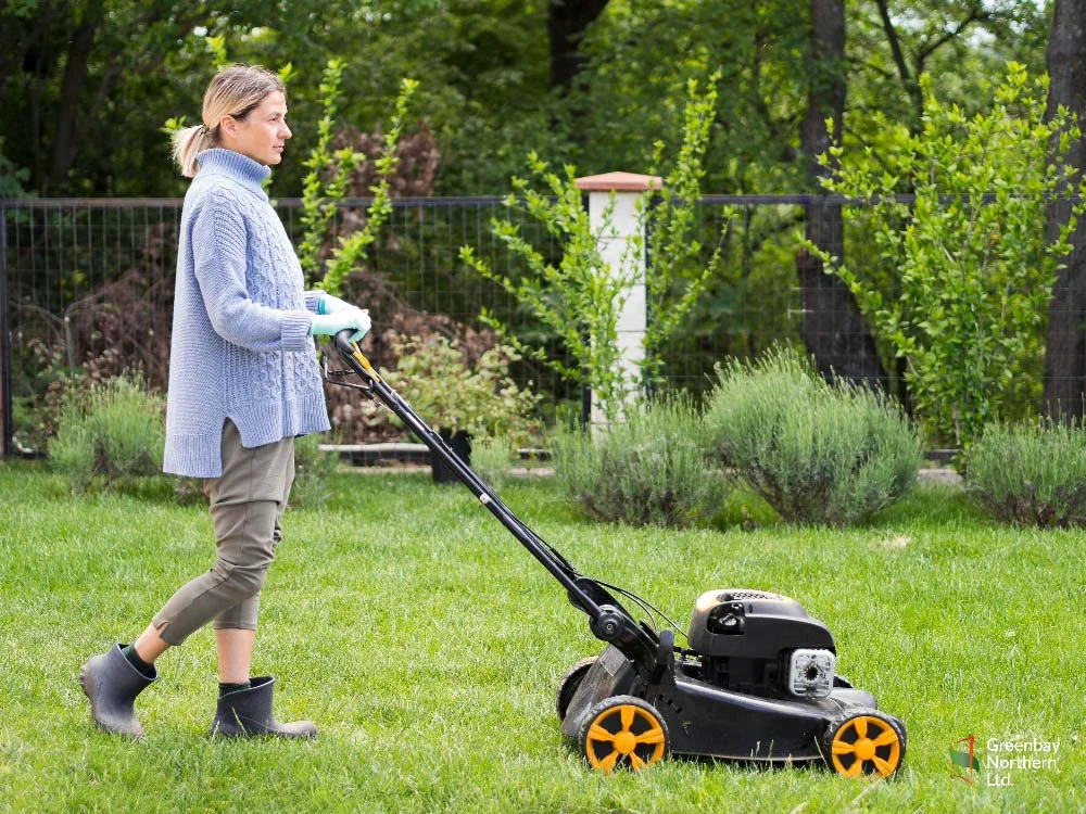Person mowing a lawn with a black lawnmower, wearing a blue sweater and boots.